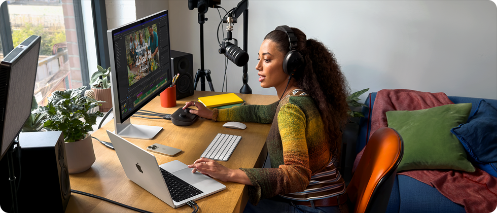 امرأة في منزلها تعمل خلف مكتبها على MacBook Pro متصل بشاشتين خارجيتين وترتدي سماعات رأس وتتحدث في الميكروفون