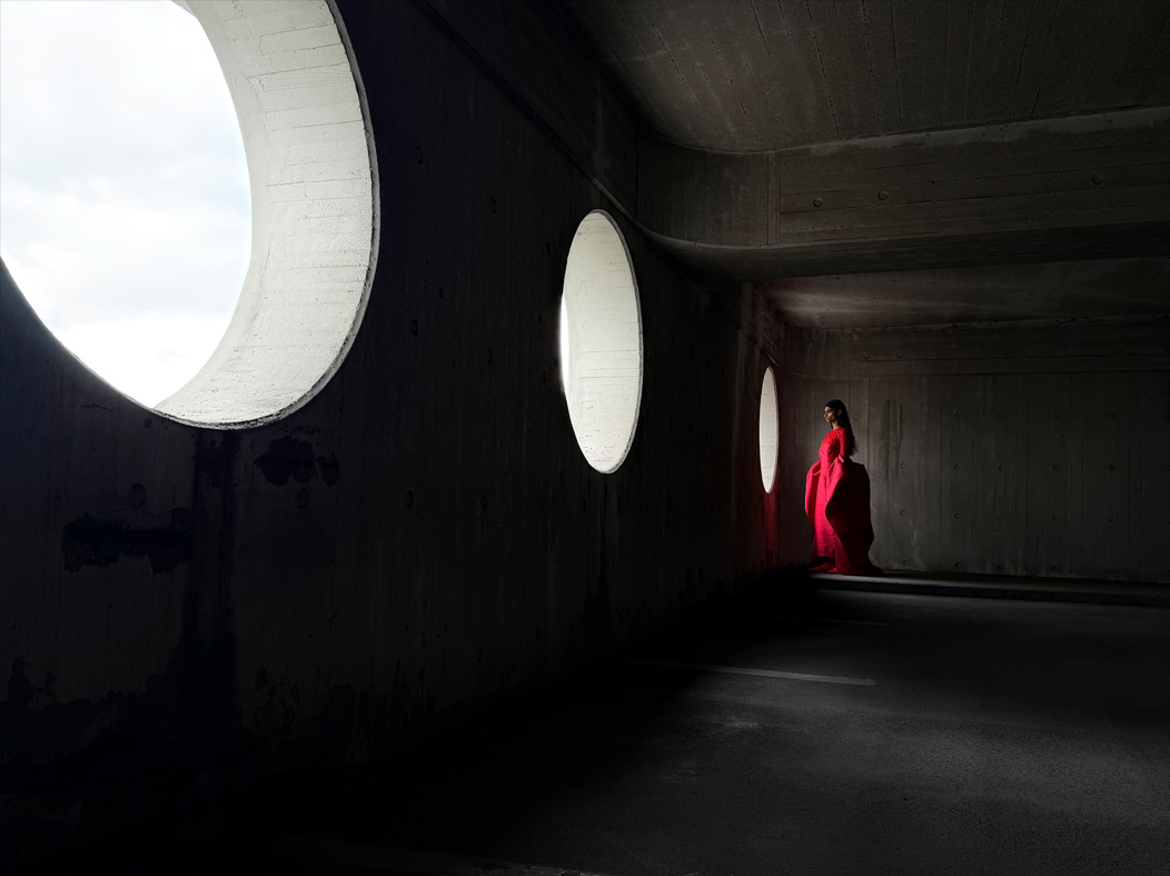 Una donna con un lungo vestito rosso fotografata in piedi in una stanza vuota. Nell’app Foto la lunghezza focale viene regolata a 28 mm, per mettere in evidenza le capacità del sistema di fotocamere Pro