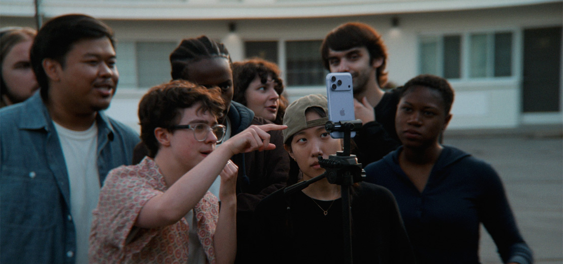 Two young filmmakers direct a scene to group of young people using an iPhone 18 Pro in Glacier color mounted on a tripod
